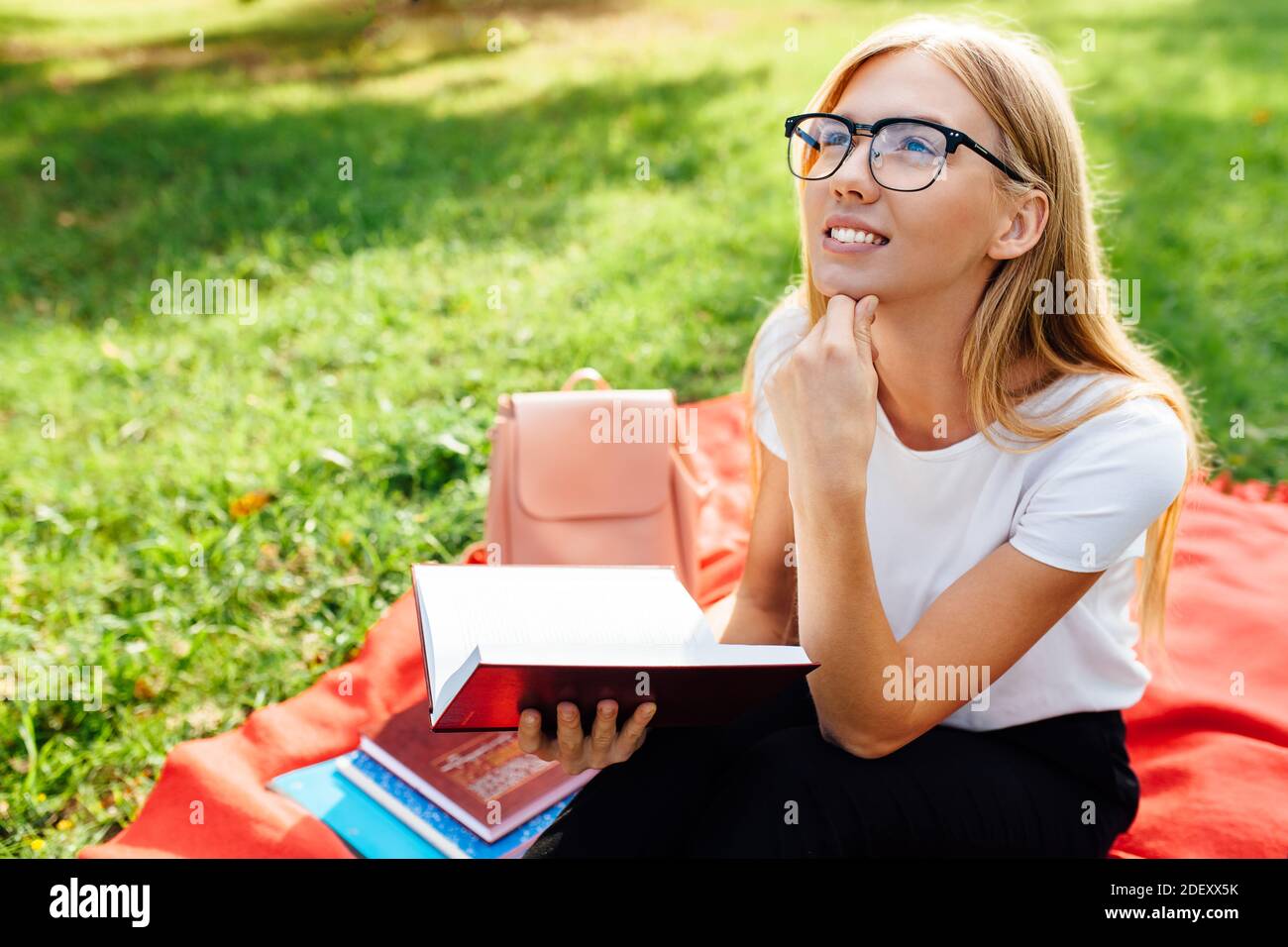 Ritratto di una bella studentessa pensante con occhiali seduti su una coperta rossa nel Parco, leggendo un libro e sognando all'aria fresca, Foto Stock