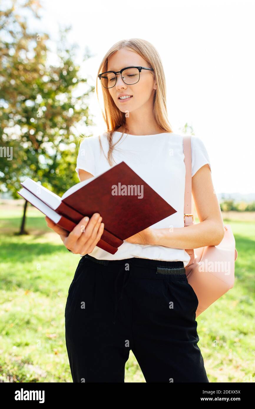 Ritratto di una bella studentessa con occhiali che legge un libro all'aria aperta, camminando nel Parco dopo la lezione Foto Stock