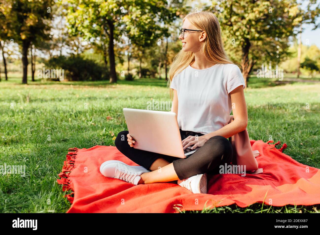 L'immagine di una bella studentessa con occhiali, che lavora su un computer portatile, seduto su una coperta rossa nel Parco facendo compiti Foto Stock