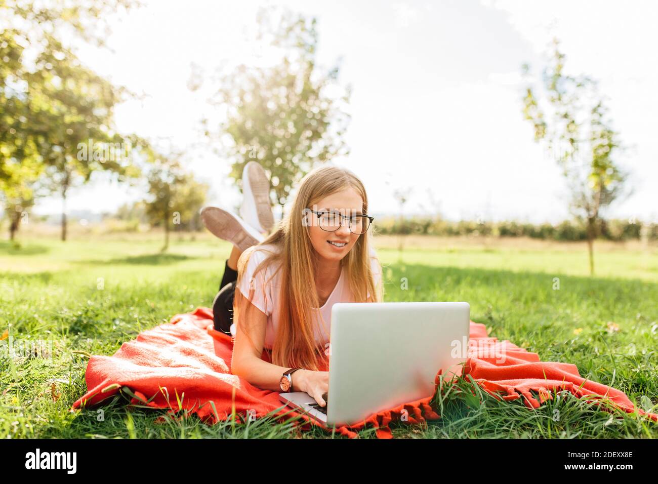 L'immagine di una bella studentessa con occhiali, che lavora su un computer portatile, seduto su una coperta rossa nel Parco facendo compiti Foto Stock