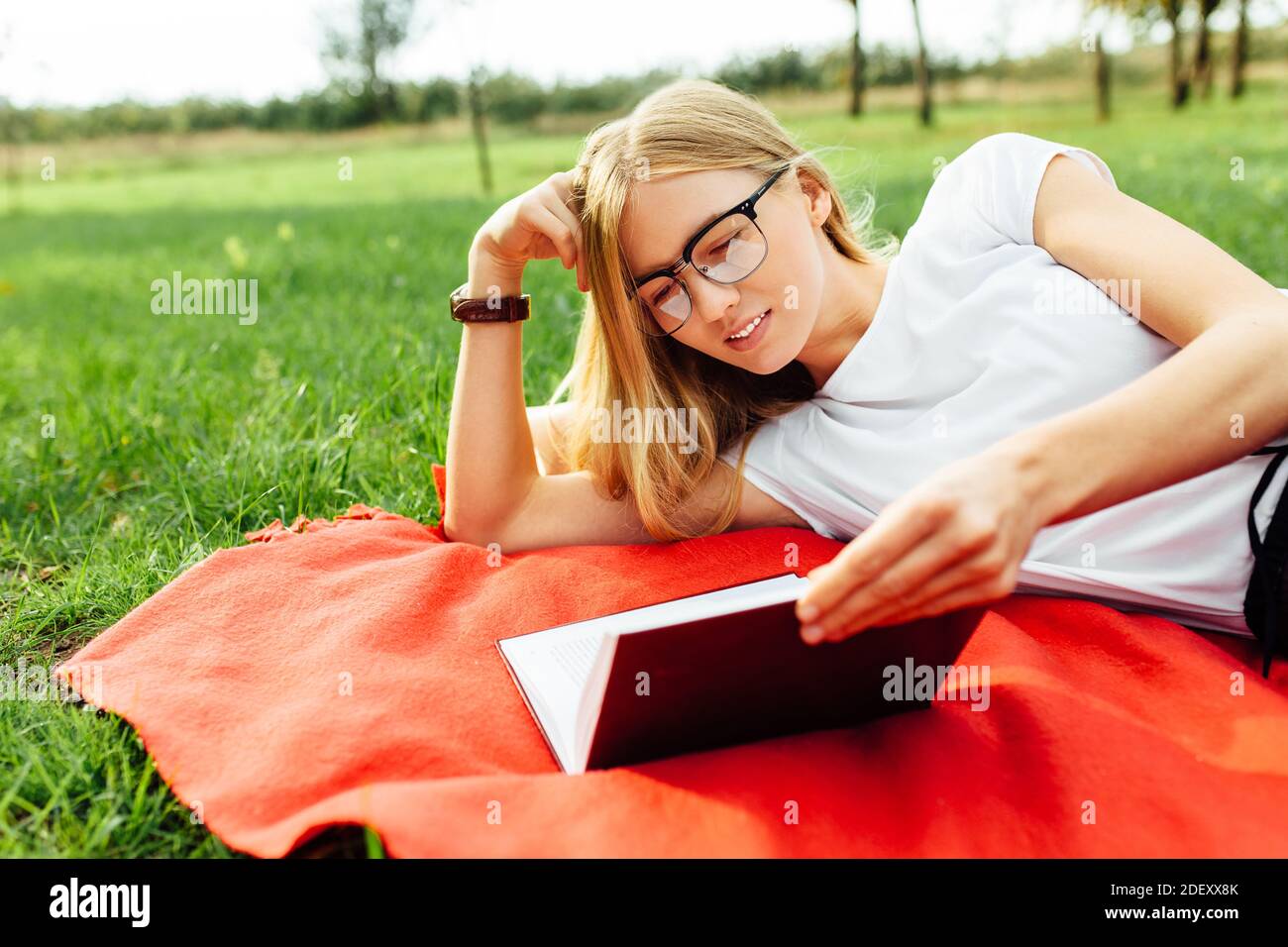 Ritratto di una bella ragazza studentessa con occhiali, seduta su una coperta rossa nel Parco, leggendo un libro all'aria fresca, riposo dopo classe Foto Stock