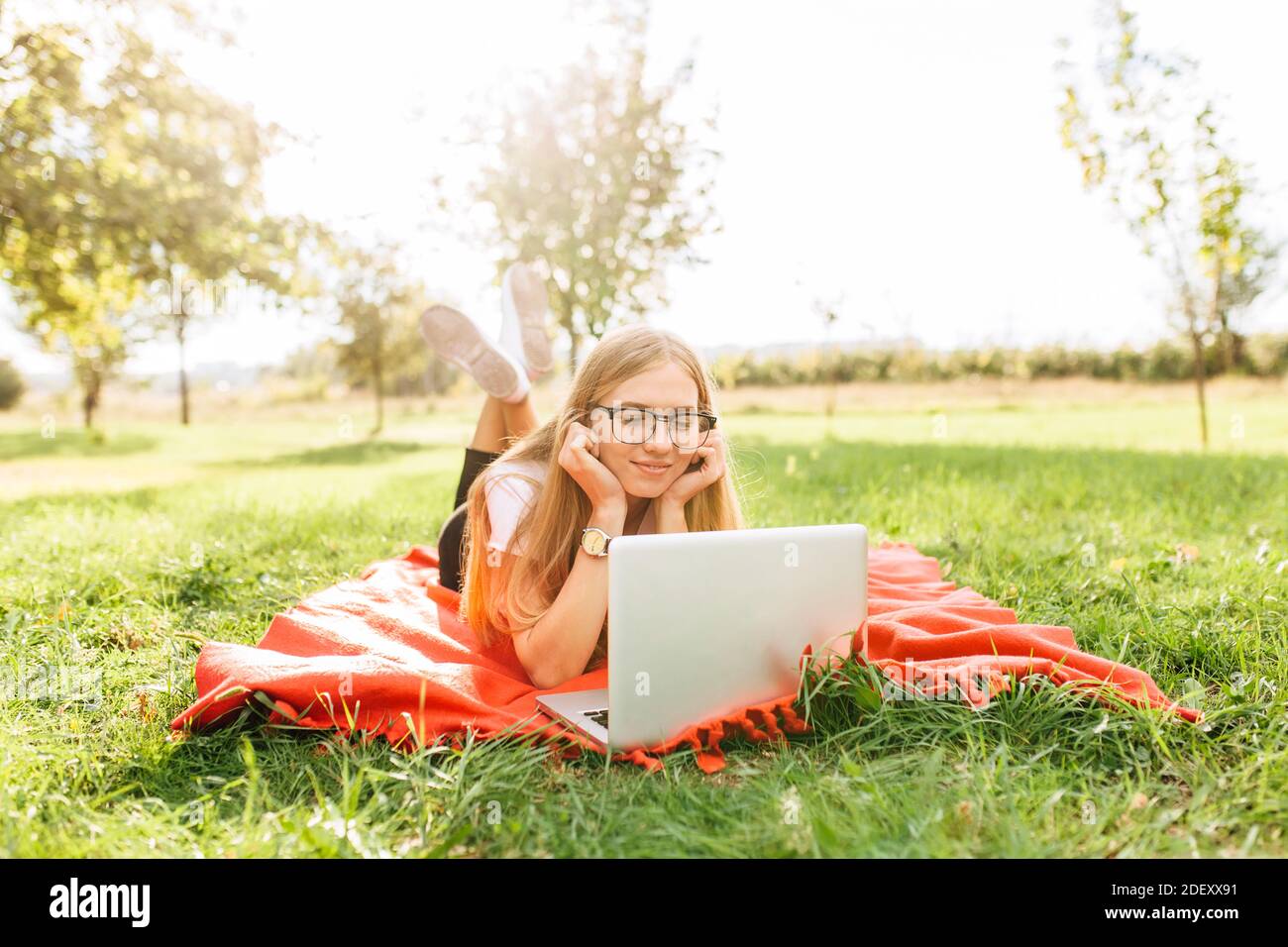 Immagine di una bella studentessa con occhiali che lavora su un computer portatile di buon umore nel Parco, sdraiato su una coperta Foto Stock