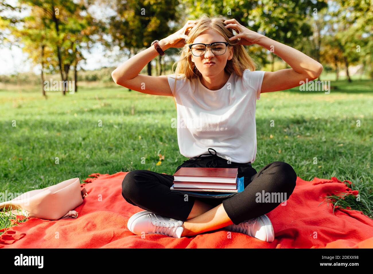 Ritratto di uno studente con occhiali stanco di studiare, seduto su una coperta rossa nel Parco con libri Foto Stock