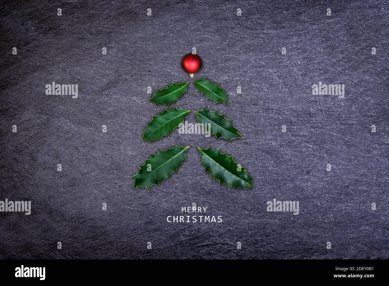 Albero di Natale fatto di foglie di agrifoglio e rosso bauble su pietra scura. Cartolina di Natale. Foto Stock