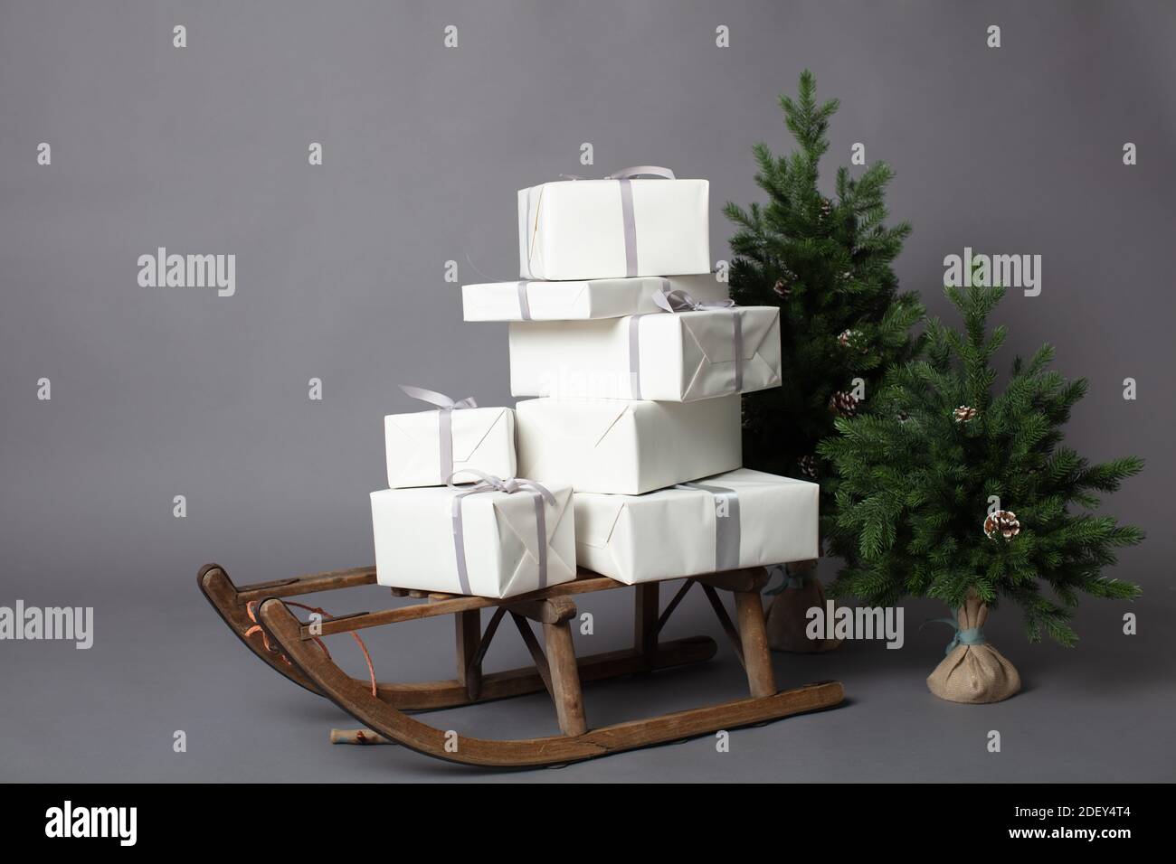 Slitta rustica con un paio di scatole regalo bianche confezionate accanto a due alberi di Natale in miniatura. Immagine orizzontale su sfondo grigio con spazio per la copia. Foto Stock