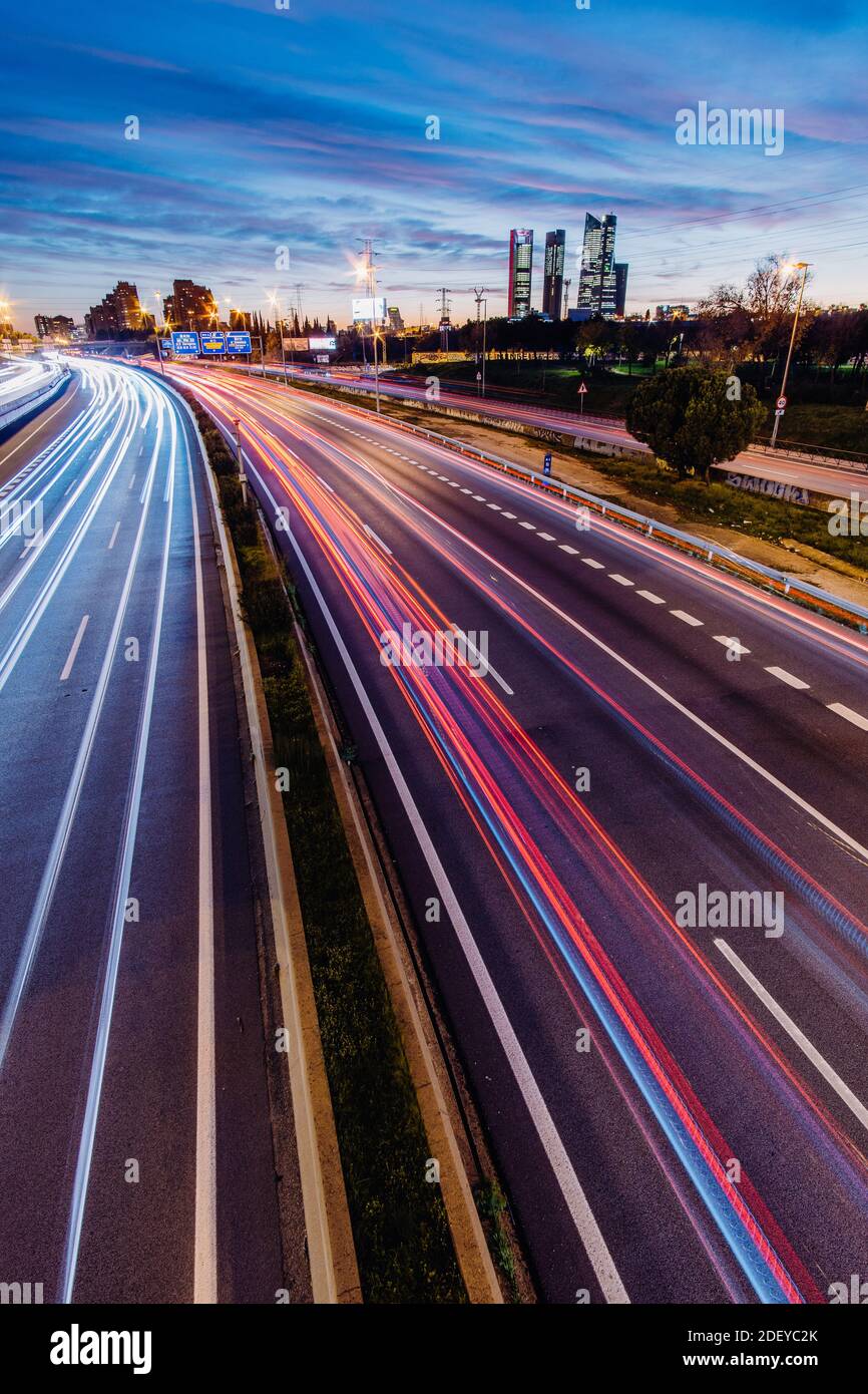 Autostrada A-1in Madrid Spagna con sentieri per auto leggere all'ora di punta al tramonto con Cuatro Torres Business Area sullo sfondo. Toni blu nel cielo in v Foto Stock