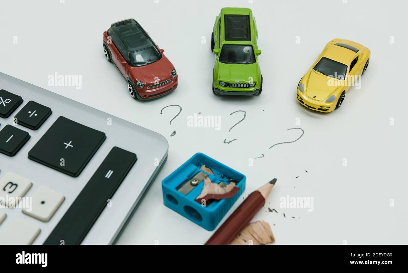 Su una superficie bianca, tre auto giocattolo, tre punti interrogativi, un affilatore a matita, la punta della matita e una calcolatrice. Foto Stock