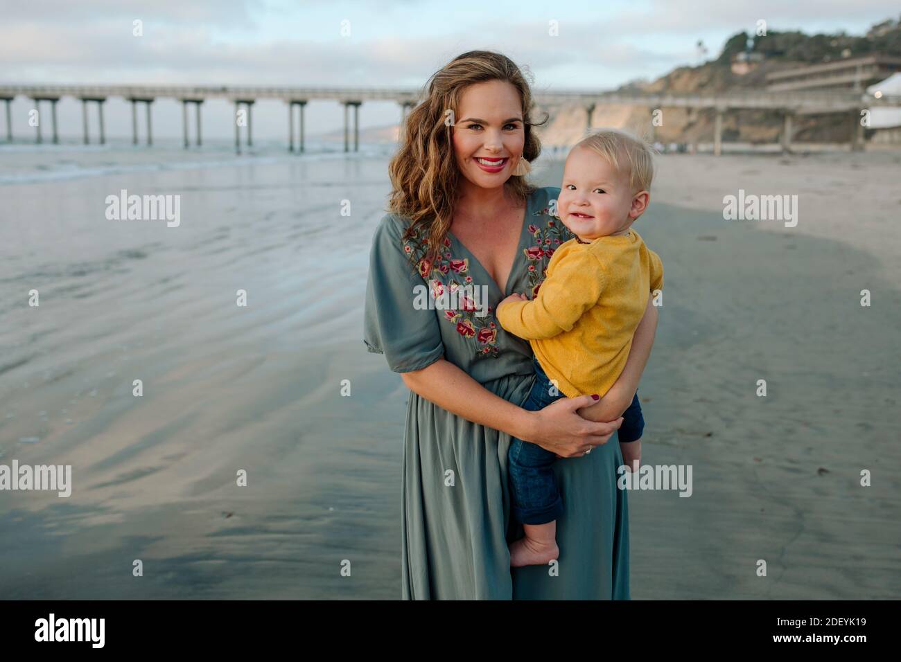 Bella mamma di 30 anni che tiene il bambino in spiaggia con molo sullo sfondo Foto Stock