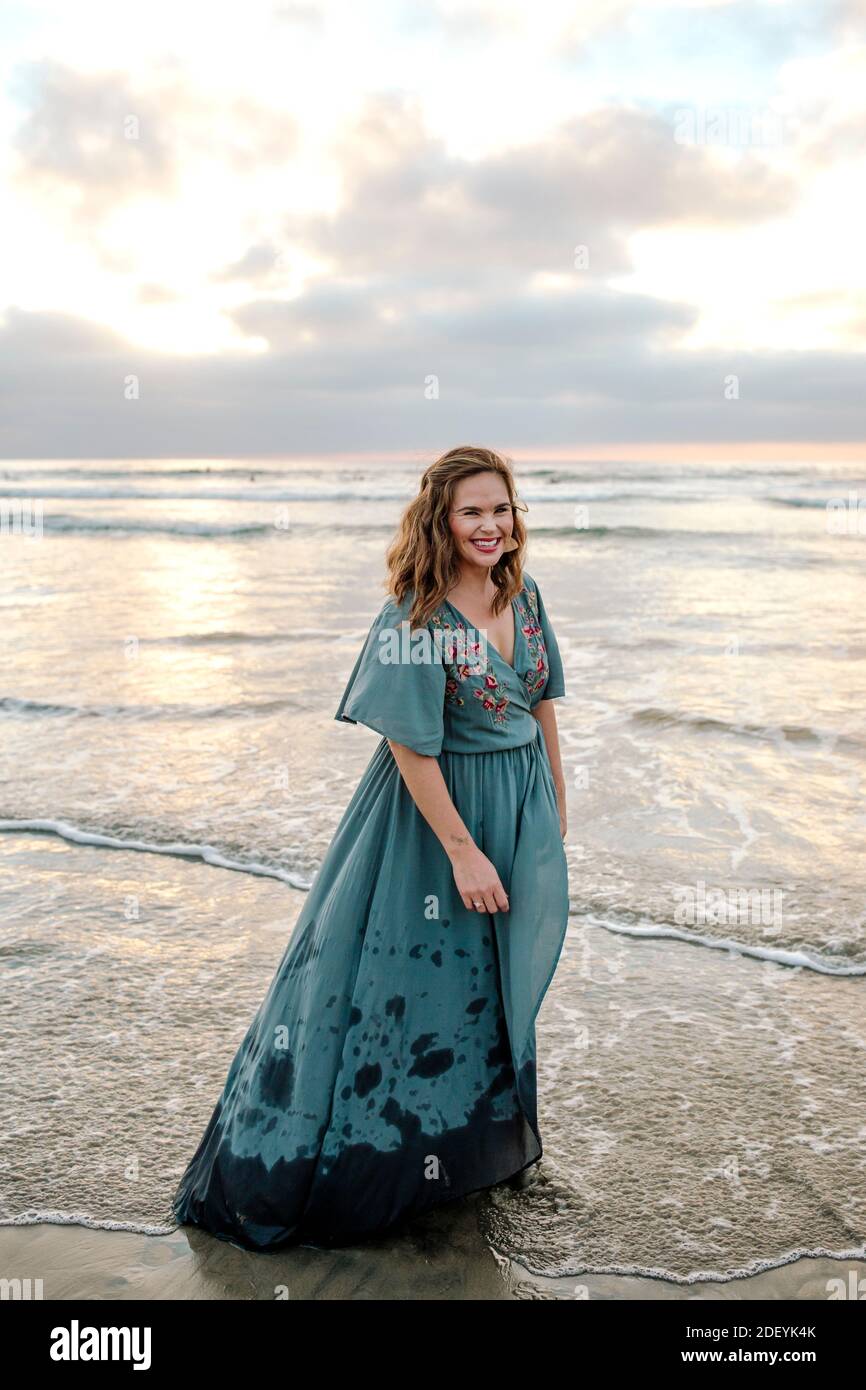 Splendida donna che indossa maxi abito Wading in mare surf al tramonto Foto Stock