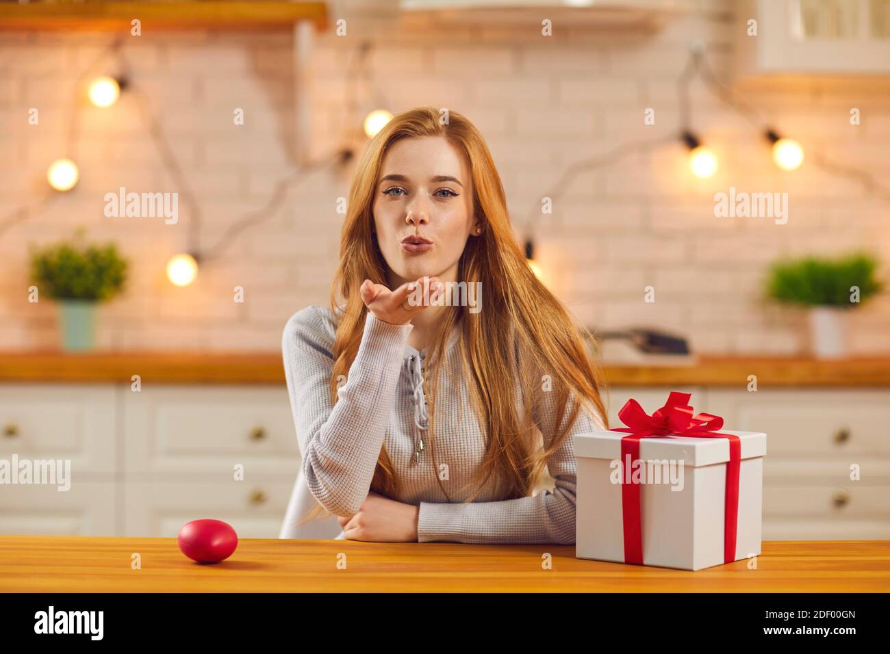 Bella giovane donna seduta al tavolo da cucina con San Valentino presente e soffiare un bacio alla macchina fotografica Foto Stock