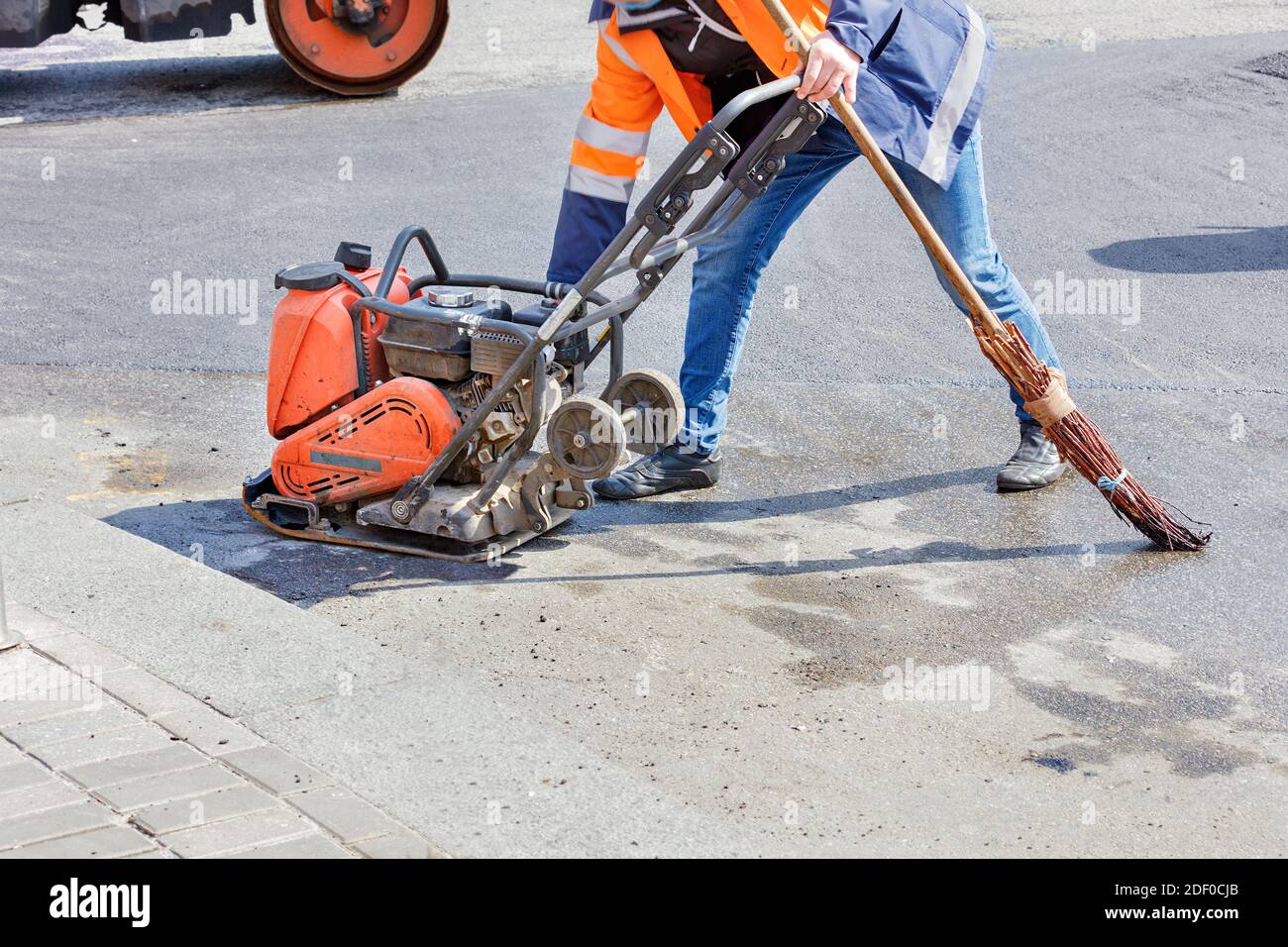 Un operatore stradale utilizza un compattatore vibrante a benzina, un rullo vibrante e una vecchia scopa per compattare l'asfalto su un tratto di strada. Foto Stock