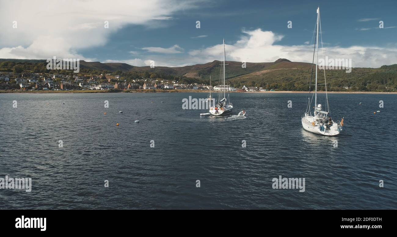 Barche sulla costa oceanica di montagne verdi isola di Arran aereo. Crociera estiva su yacht di lusso a vela con passeggeri al porto di Brodick. Baia del mare della Scozia con trasporto dell'acqua al colpo cinematografico del drone Foto Stock