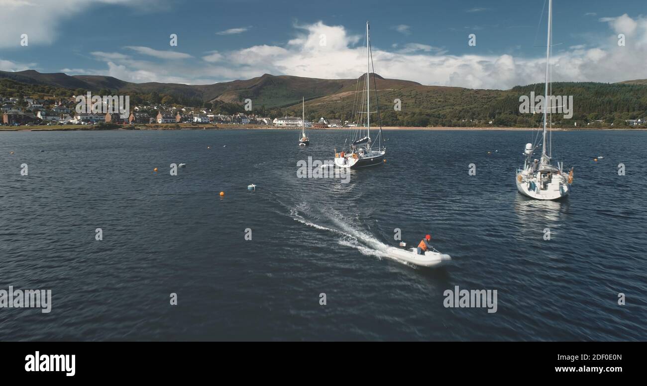 Barche a motore e barche a motore a mare baia di verde isola costa aerea. Paesaggio montano a Bridick città molo, Arran isola, Scozia. Crociera estiva per passeggeri su barche a vela, motoscafi sulla verde costa collinare Foto Stock