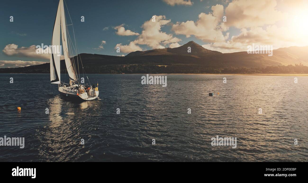 Sun barca a vela all'antenna di riflessione dell'acqua della baia dell'oceano. Crociera estiva sull'isola delle montagne verdi di Arran su yacht di lusso con passeggeri. Spettacolare costa marina scozzese con nave Foto Stock