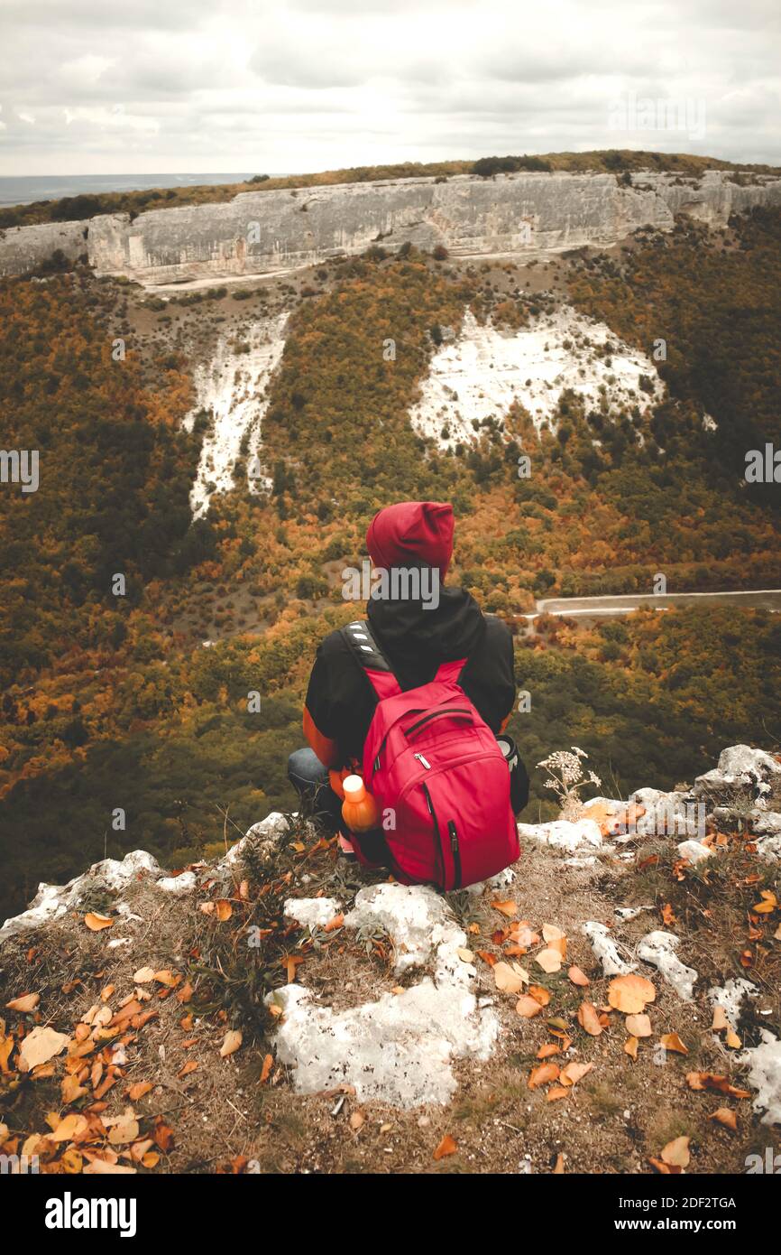 Il viaggiatore si siede sul bordo con lo zaino rosso e la bania, rilassarsi e godersi il paesaggio. Panorama verticale della foresta di montagna d'autunno Foto Stock
