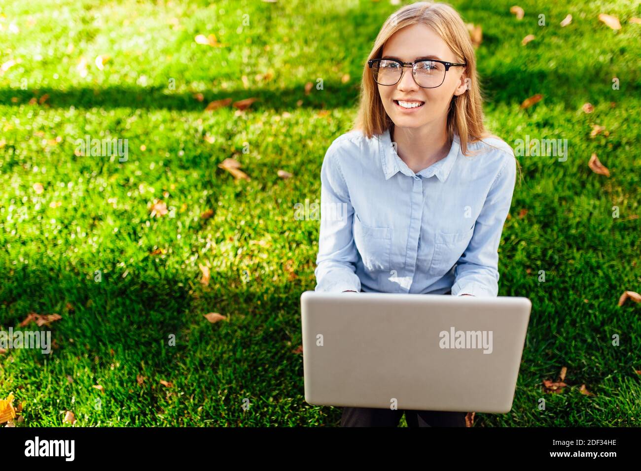 Giovane ragazza attraente con occhiali, Donna seduta sull'erba, lavorando su un computer portatile, in un parco cittadino su un prato verde all'aperto. Business conce freelance Foto Stock