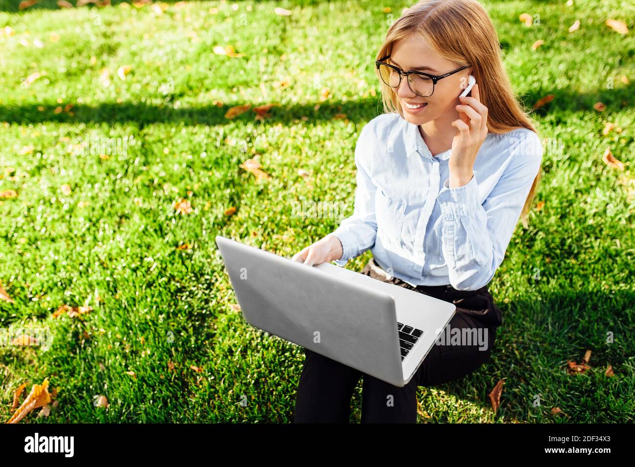 Ritratto di una giovane donna attraente che indossa occhiali seduti su erba verde nel parco in una giornata calda, lavorando con un computer portatile e cuffie wireless Foto Stock