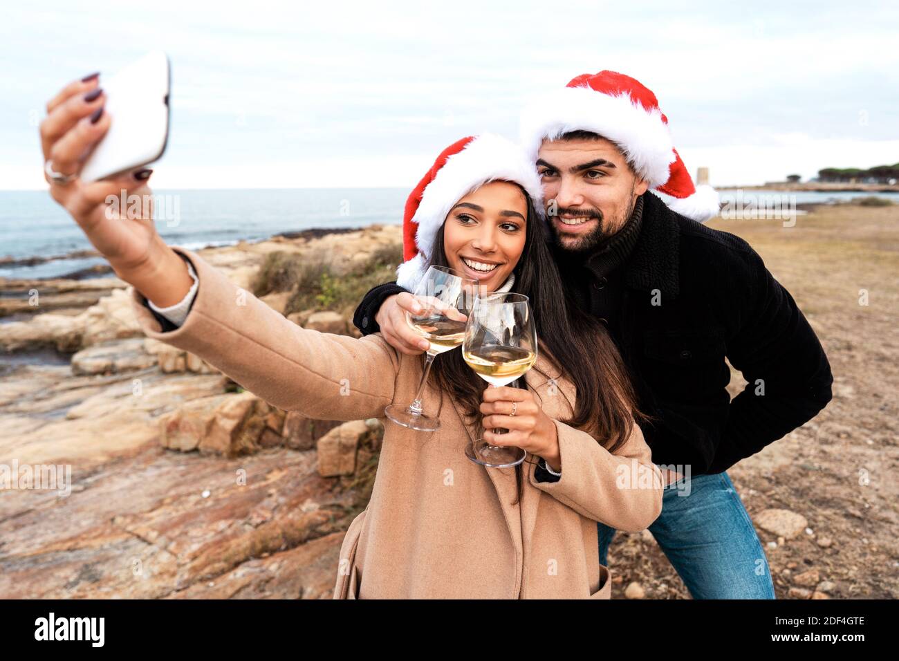 Giovane bella coppia multirazziale che fa un autoritratto all'aperto dentro Resort marittimo con cappello Babbo Natale che tiene bicchieri di vino bianco Celebrazione di Christm Foto Stock