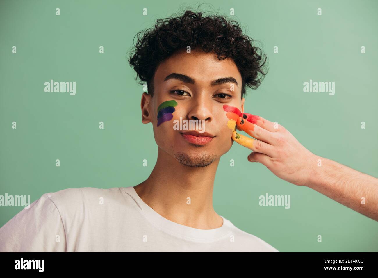 Ritratto di giovane uomo con volto dipinto con bandiera arcobaleno di orgoglio gay. Pittura a mano lgbt colore arcobaleno sul viso di un uomo. Foto Stock