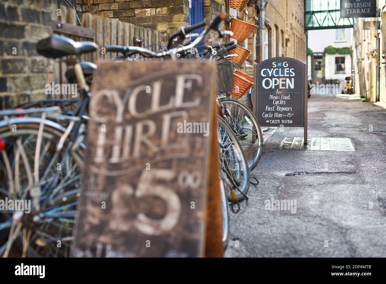 Strade acciottolate a Cambridge, Regno Unito, con cartelli per il noleggio di biciclette e turisti e locali che si recano per la loro attività quotidiana in bicicletta Foto Stock