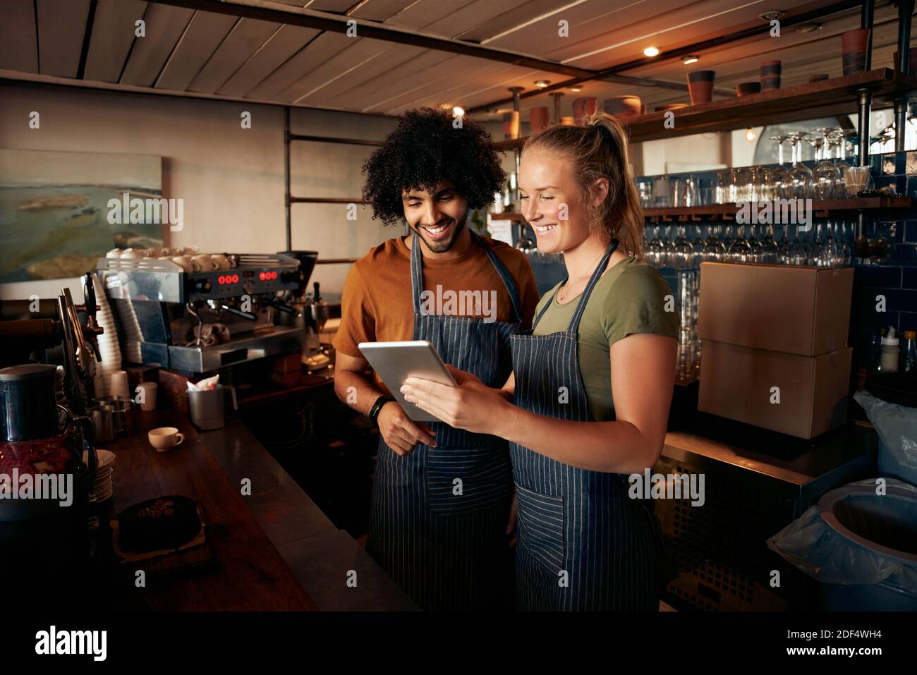 Cameriere sorridente e cameriera con grembiule e tablet digitale in piedi dietro il banco del caffè Foto Stock