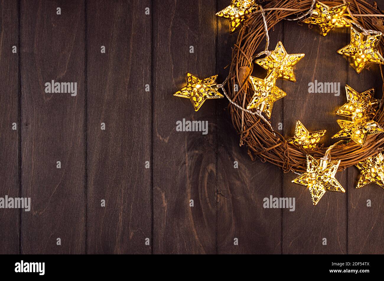 Ricco sfondo festivo - corona di natale, stelle dorate con glitter garland incandescente luce calda su tavola di legno marrone scuro, vista dall'alto, bordo. Foto Stock