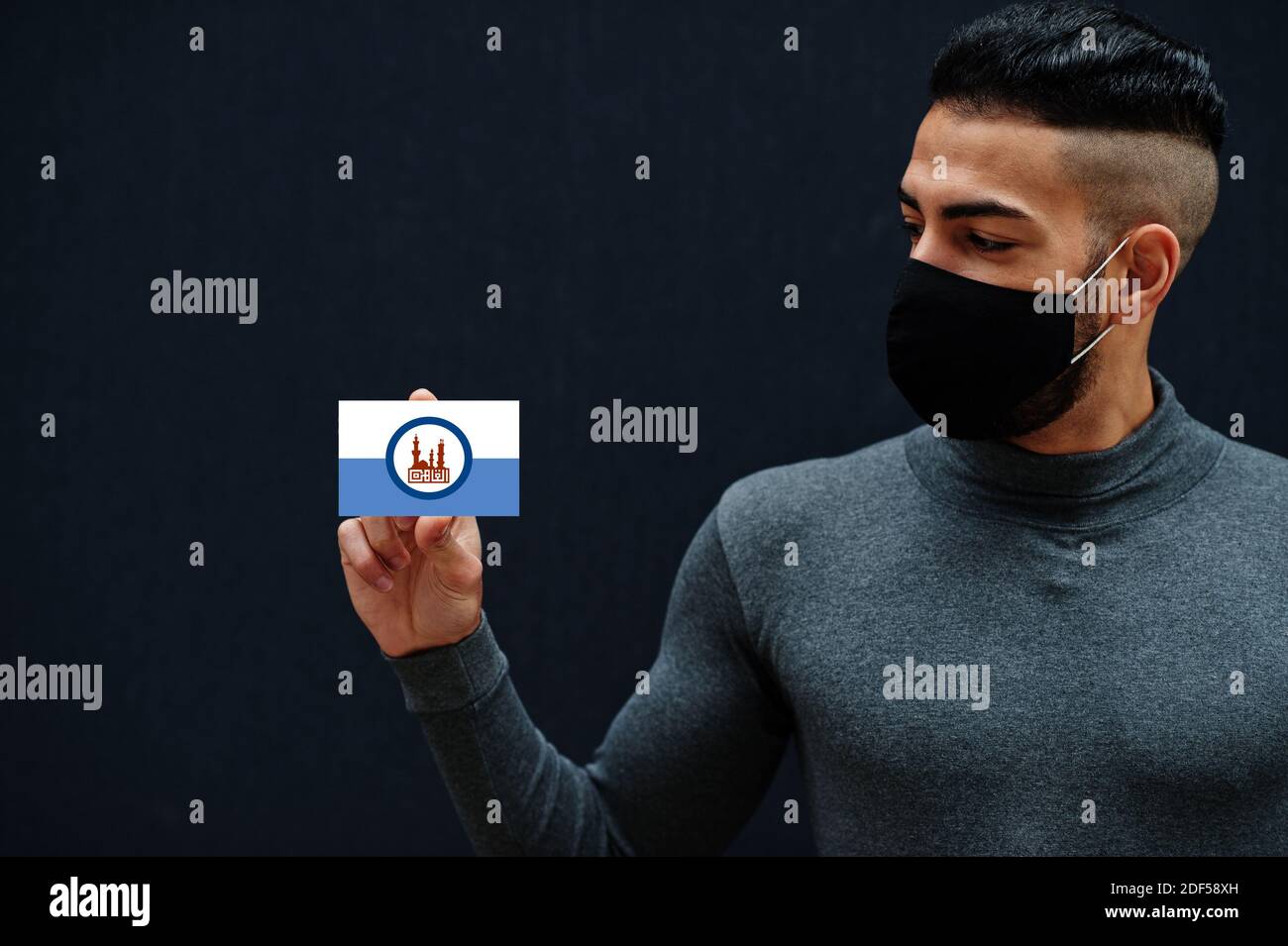 L'uomo egiziano in tleneck grigio e maschera di protezione viso nero mostra Cairo bandiera sfondo isolato. Governatorati di Egitto coronavirus concetto. Foto Stock