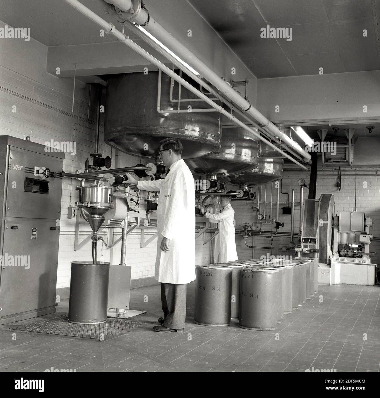 Anni '1950, storici tecnici con rivestimento bianco che lavorano in un'area di produzione sotto grandi fusti di metallo presso la fabbrica chimica Sandoz, dove vengono prodotti coloranti sintetici per abbigliamento e cibo, Inghilterra, Regno Unito. Foto Stock