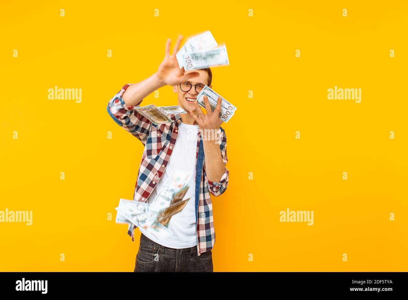 Ritratto di un uomo felice, indossando occhiali e una camicia di plaid, gettando le banconote di denaro su uno sfondo giallo Foto Stock