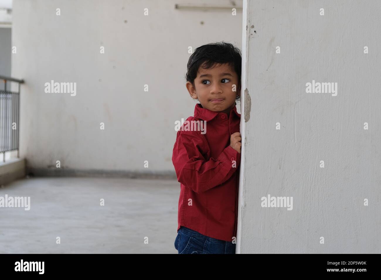 Ritratto di indiano carino bruna allegro ragazzo Tamil bambino indossare una camicia rossa vivace mentre ci si trova su un balcone uno sfondo urbano bianco Foto Stock