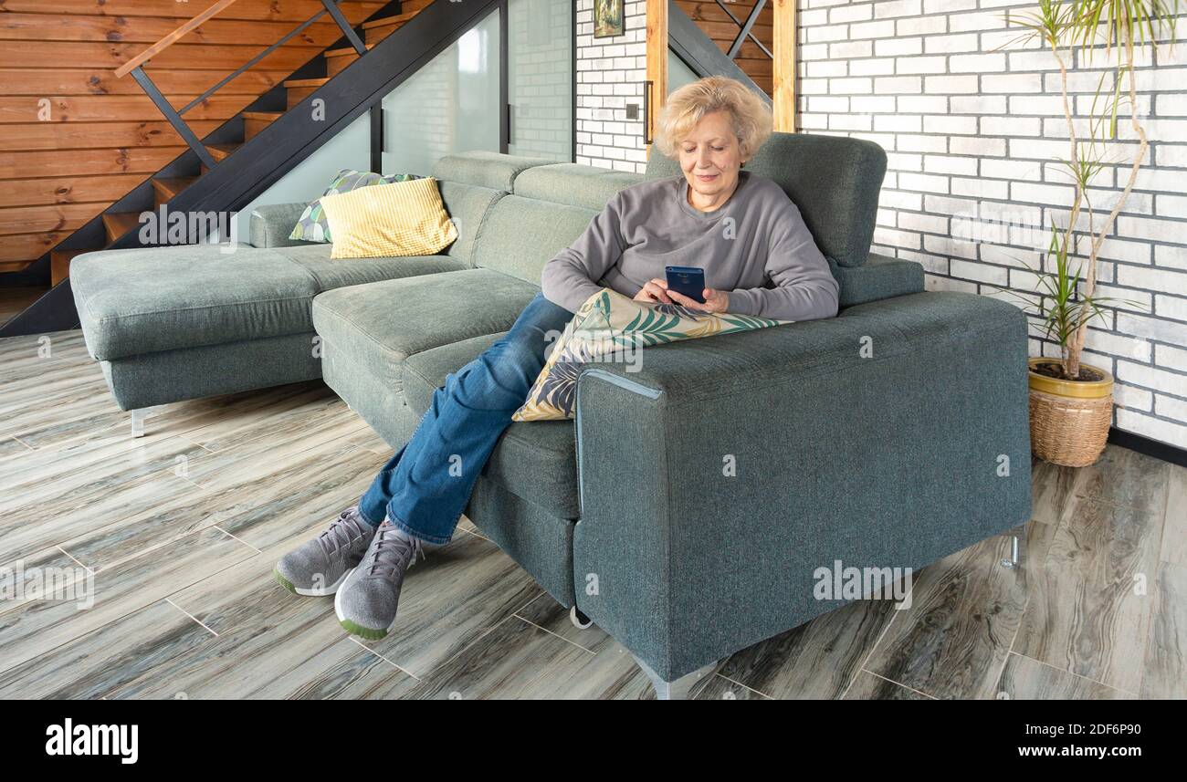 Una signora anziana adulta effettua pagamenti via internet mentre si siede sul divano a casa nel soggiorno. Comunicazione remota su social network e messe Foto Stock