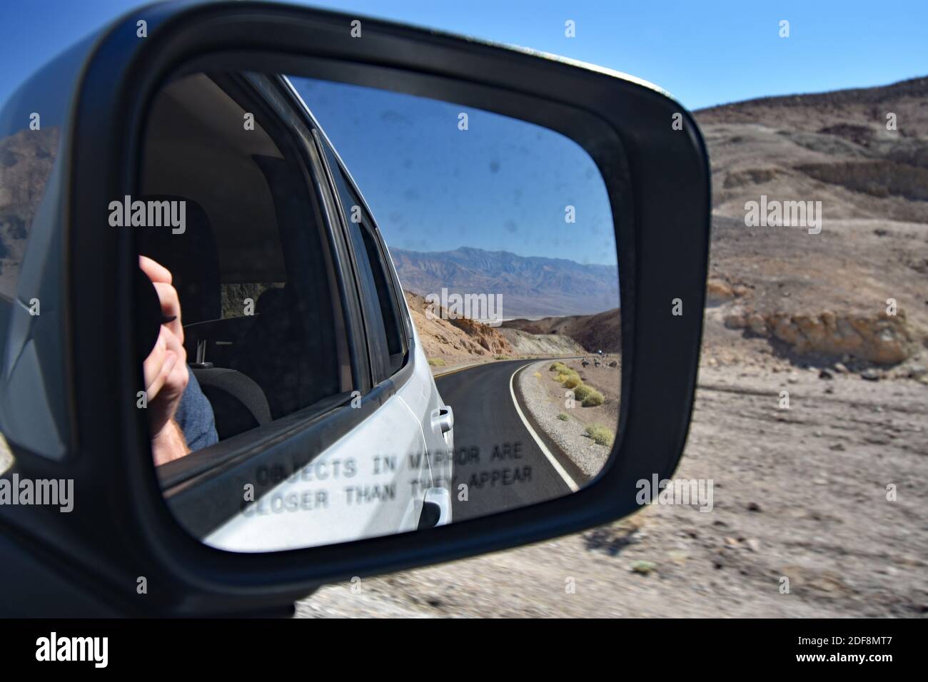 Un maschio scatta una foto dello specchio dell'auto mentre si trova lungo Artists Drive nel Death Valley National Park, USA. La fotocamera, le mani e le catene montuose vengono riflesse. Foto Stock