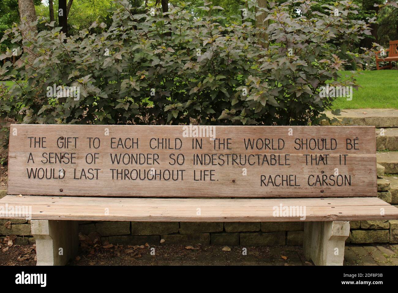 Una panca di parco di legno incisa con una citazione ispiratrice lungo un muro di fermo di pietra, arbusti, alberi ai giardini botanici rotativi a Janesville, Wisconsin, Foto Stock
