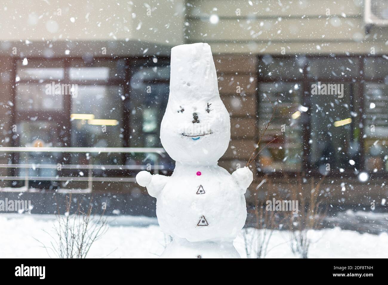 Grande bianco sorridente surgelato da solo snowman stand sulla strada della città alla tempesta di neve Blizzard contro l'edificio di appartamenti o uffici sullo sfondo. Inverno panoramico Foto Stock