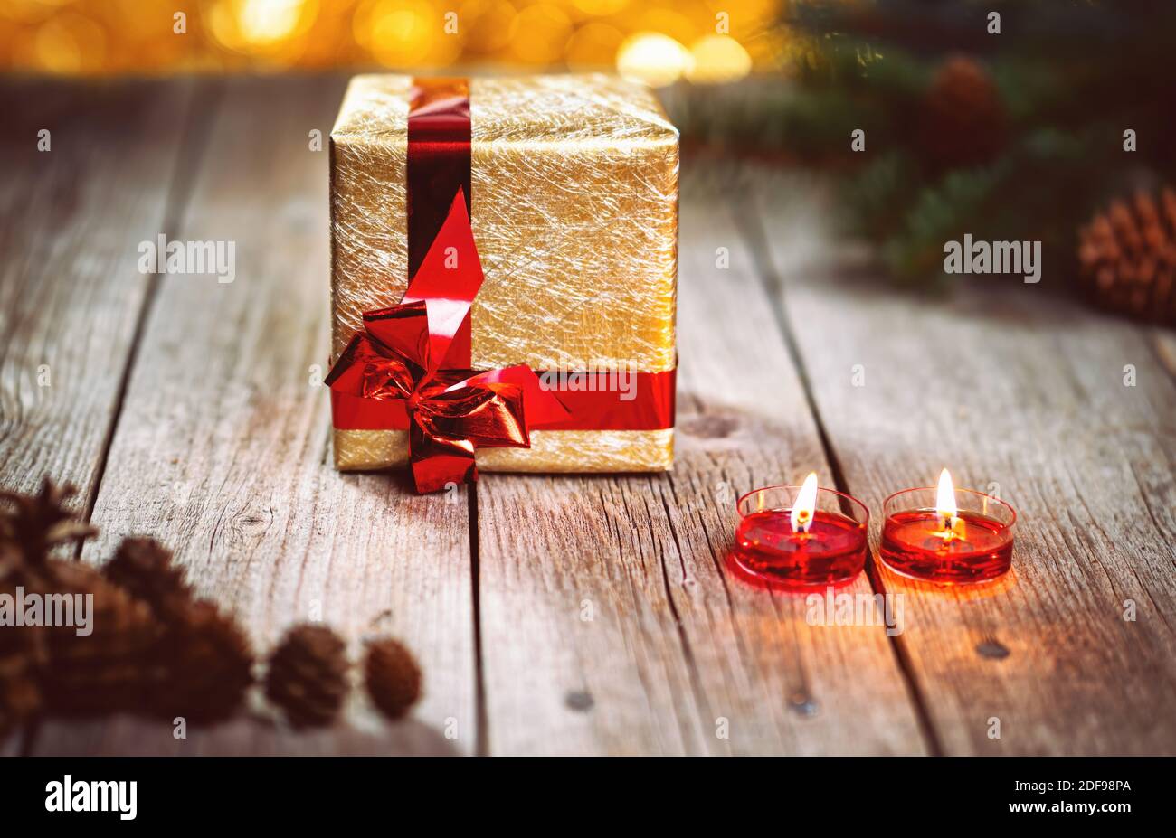 Scatola regalo d'oro e ramo di pino con coni con candele su tavolo di legno. Sfondo bokeh luci gialle. Vacanze di Natale e Capodanno a tema decoratio Foto Stock