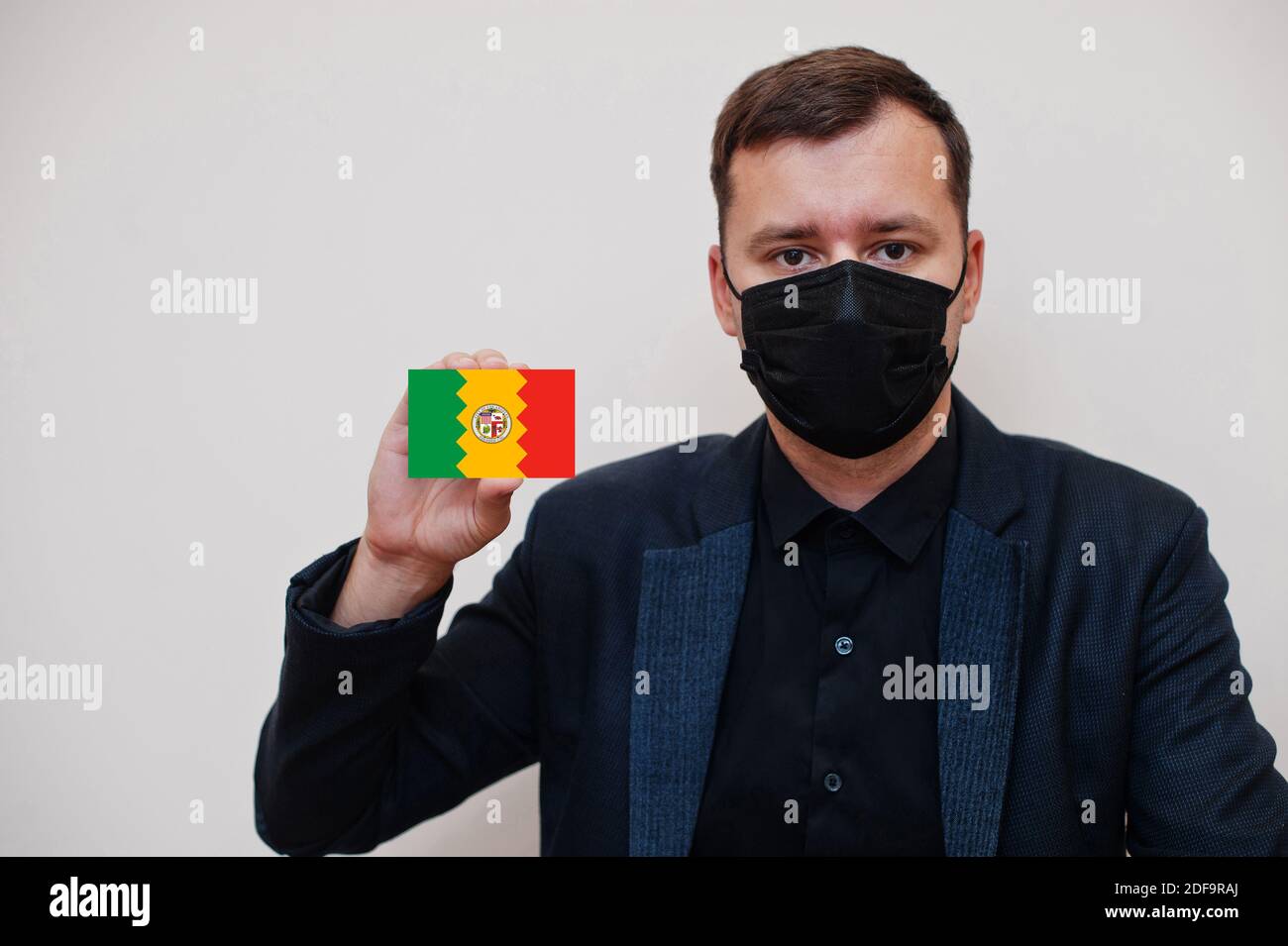 Uomo americano indossare nero formale e proteggere maschera viso, tenere Los Angeles carta bandiera isolato su sfondo bianco. Concetto di Coronavirus Covid delle città degli Stati Uniti. Foto Stock