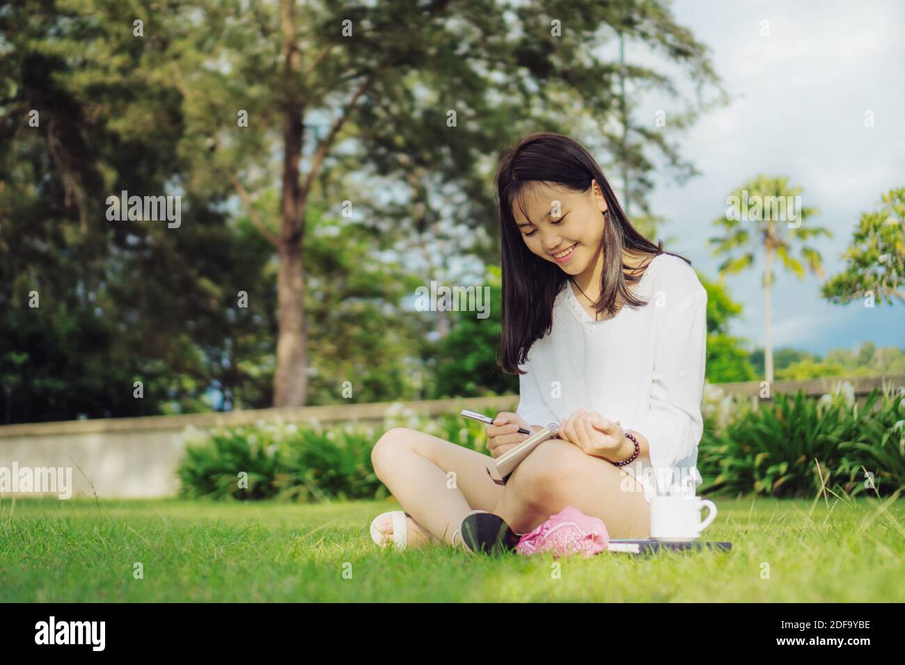 Sorridente giovane donna asiatica seduta con le gambe si incrociano su campo d'erba in un parco pubblico mentre scrive su diario. Fotografia stile di vita all'aperto. Foto Stock