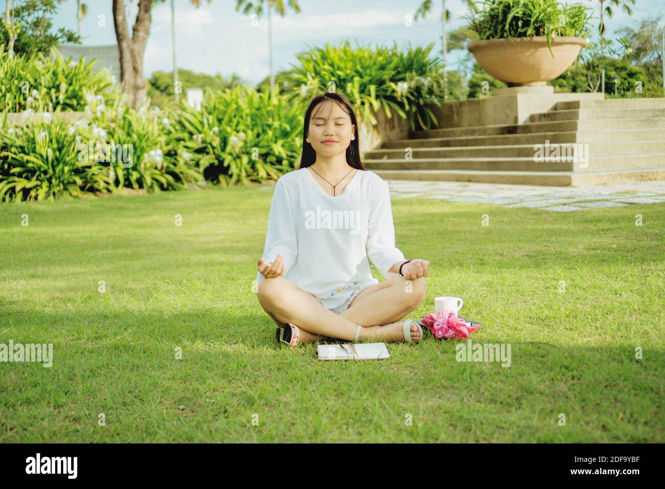 Giovane donna asiatica seduta con gambe incrociate sull'erba in un parco pubblico e meditating. Fotografia stile di vita all'aperto. Foto Stock