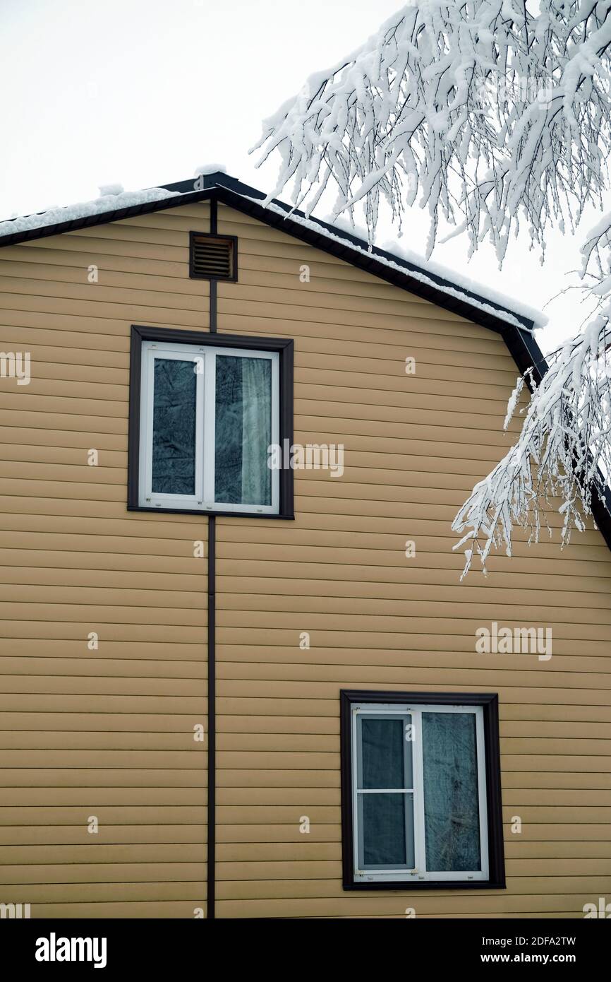Piano superiore con finestra in plastica in campagna muro coperto con fiancate beige e tetto in metallo marrone con superficie innevata e rami di albero coperti con Foto Stock
