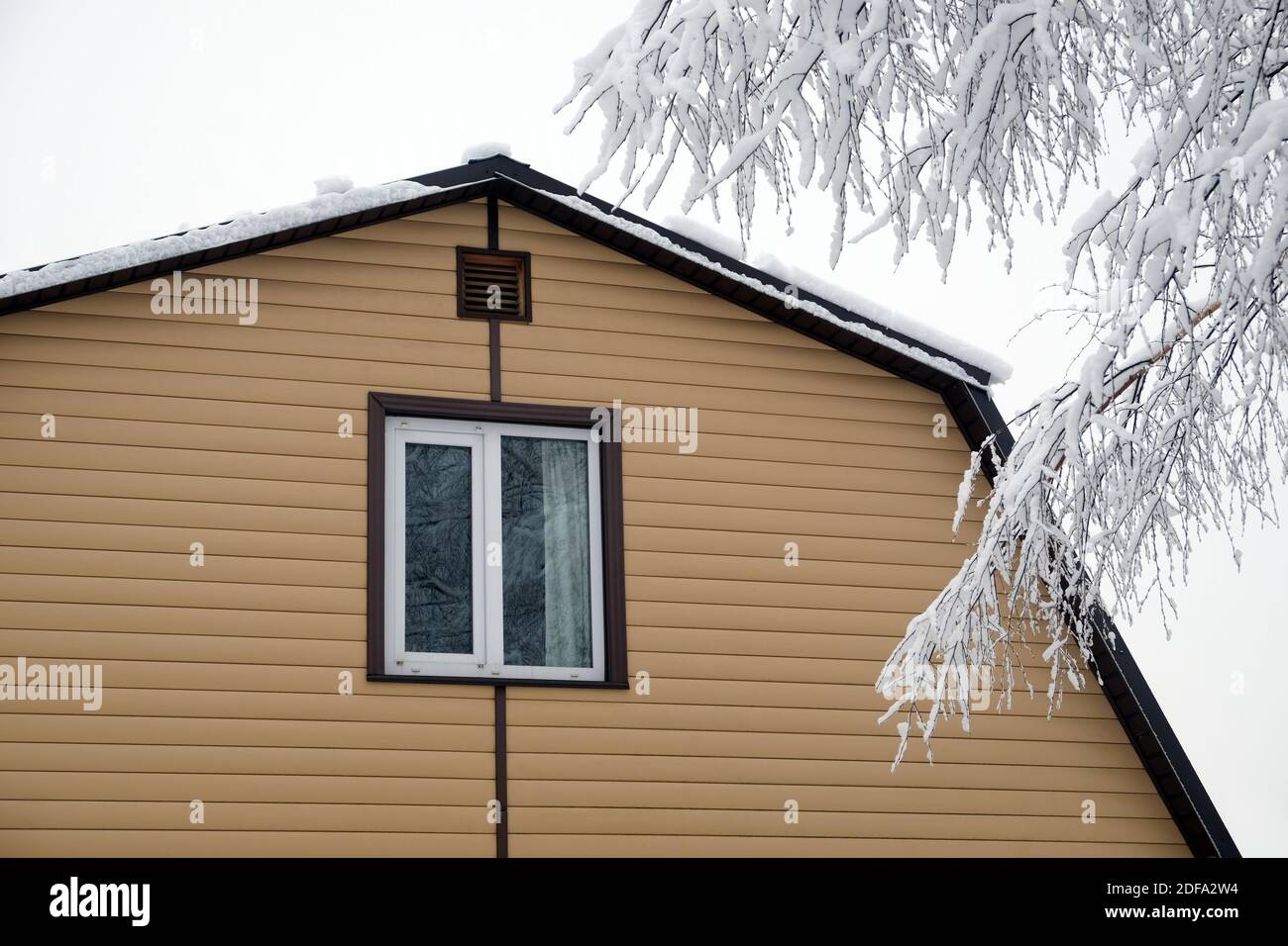 Piano superiore con finestra in plastica in campagna muro coperto con fiancate beige e tetto in metallo marrone con superficie innevata e rami di albero coperti con Foto Stock