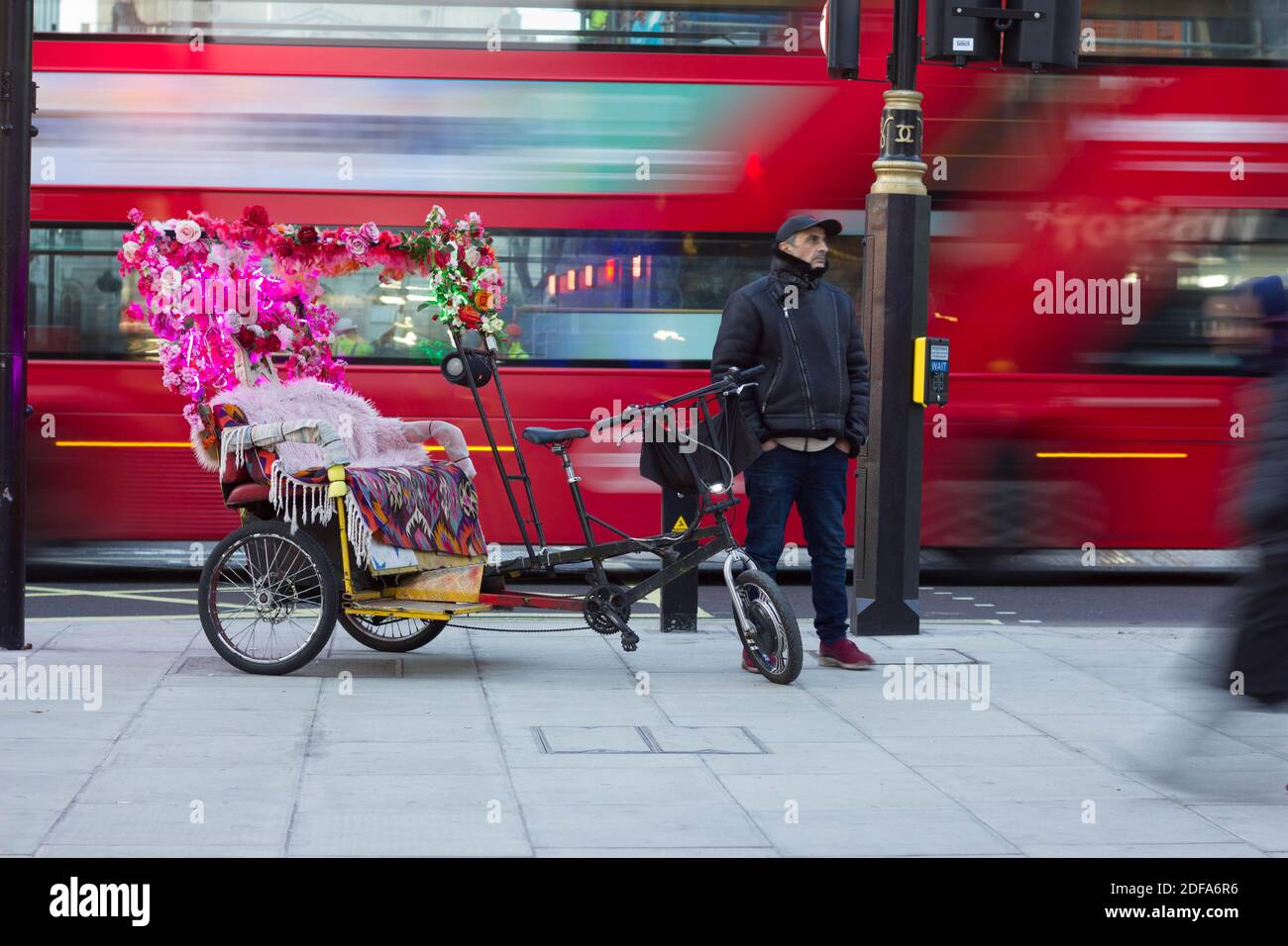 Un rickshaw, o jinrikisha, decorato con fiore, driver maschile in giacca di pelle e cappuccio di base palla in attesa di passeggero sul West End di Londra, Inghilterra Foto Stock