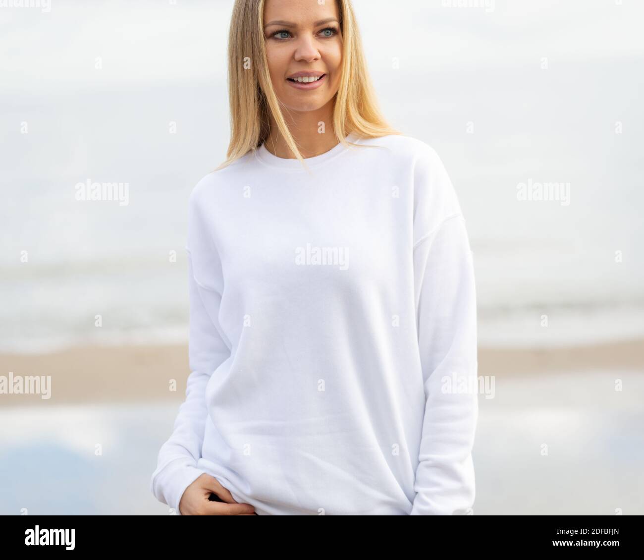 Moda mockup con spazio di copia. Donna blonde sorridente si trova in blusa bianca di fronte alla fotocamera. Vista esterna sulla spiaggia. Foto Stock