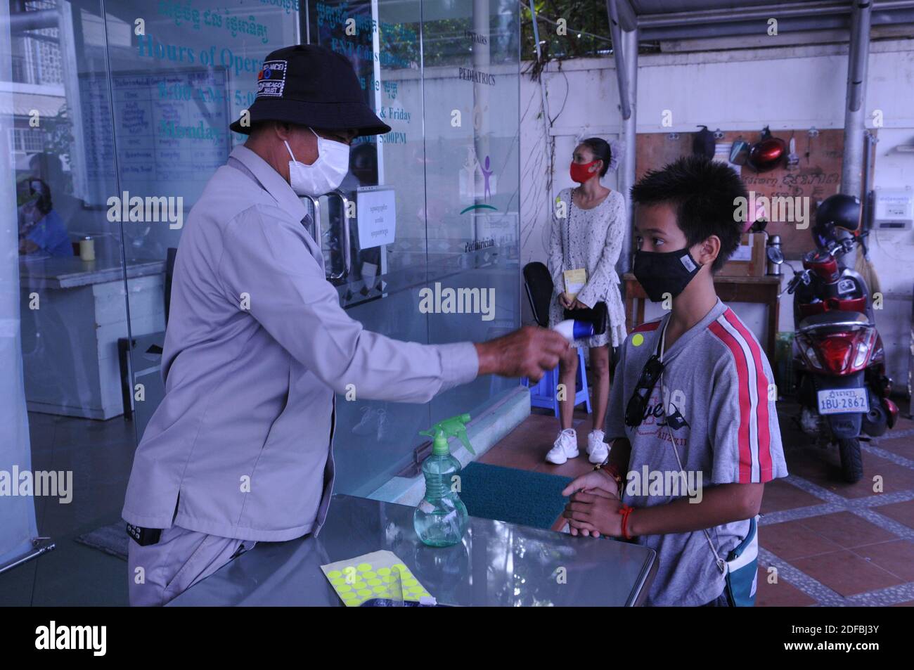 Un recente focolaio di COVID - 19 a Phnom Penh è in corso test di massa. Una razza mista (cambogiana - americana) ragazzo adolescente, con uno sguardo preoccupato sul suo viso e indossando una maschera protettiva / copertura, ottiene la sua temperatura controllata da una guardia di sicurezza all'ingresso di una clinica medica durante la pandemia di coronavirus. Phnom Penh, Cambogia. 1st dicembre 2020. © Kraig Lieb Foto Stock