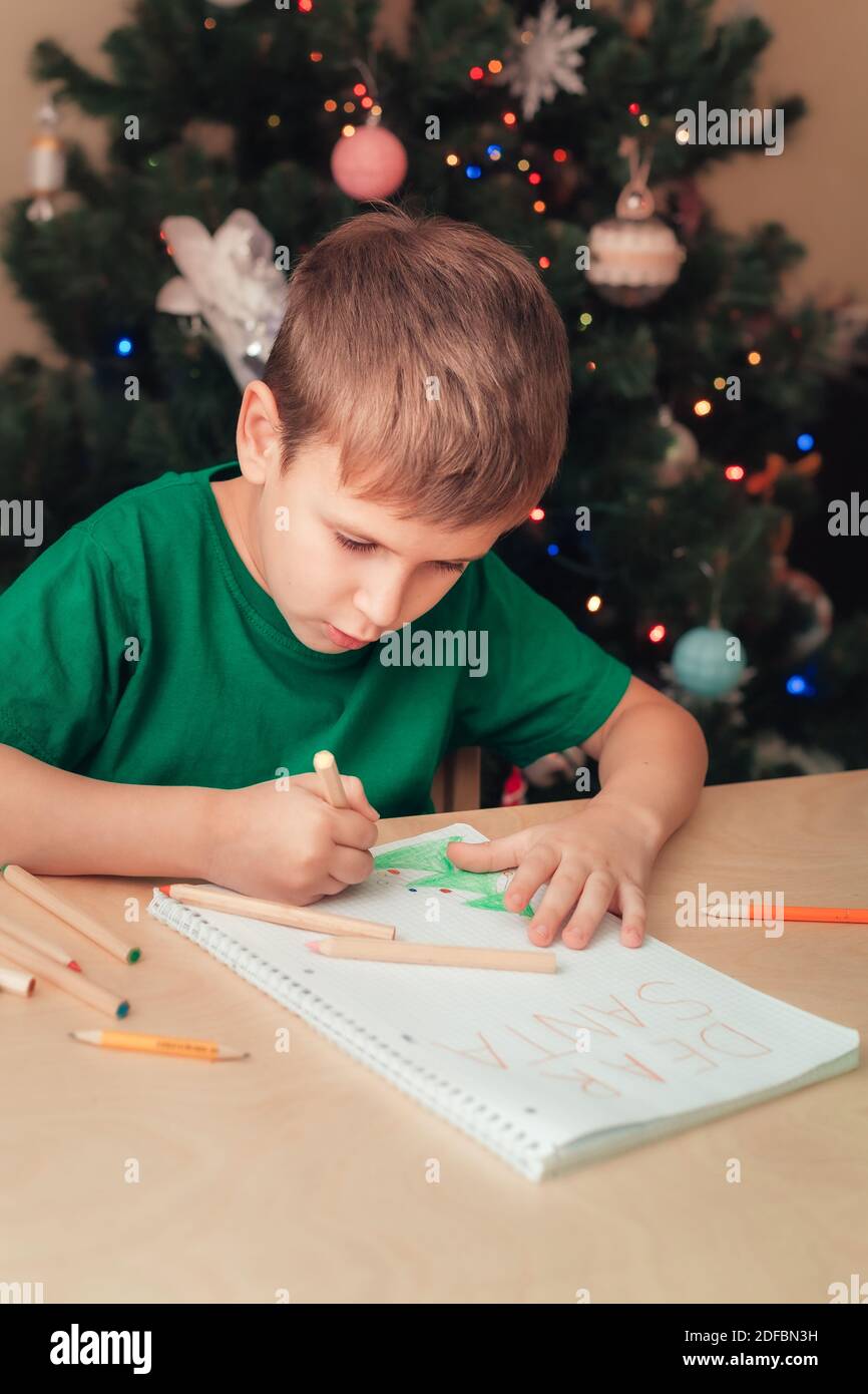 Sorridente ragazzo di 7 anni che indossa verde t-short scrivere lettera a Santa seduta dalla scrivania, albero di Natale su sfondo Foto Stock