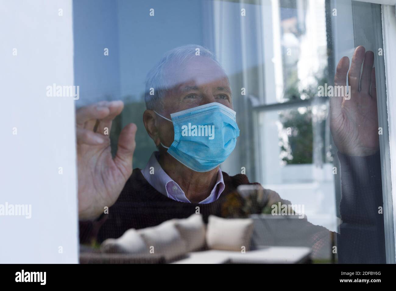 Uomo caucasico senior che indossa una maschera facciale che guarda attraverso la finestra di vetro a casa Foto Stock