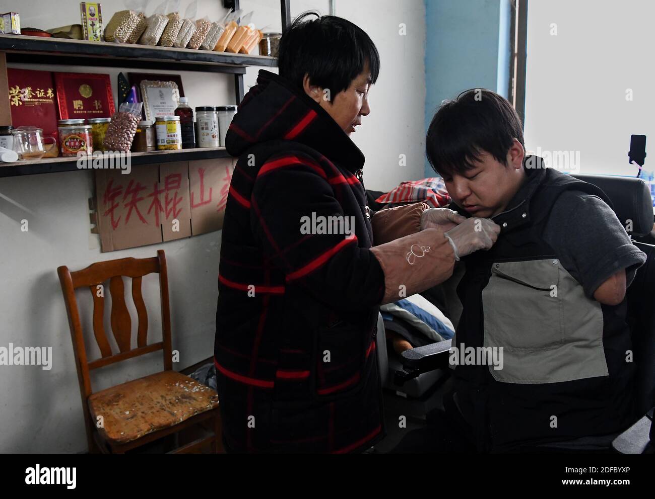 (201204) -- ZHENGZHOU, 4 dicembre 2020 (Xinhua) -- la madre di Sun Yahui lo aiuta a vestirsi a casa nel villaggio di Yangzhuang a Xinxiang, provincia di Henan della Cina centrale, 2 dicembre 2020. "Ciao, tutti! Oggi vi mostrerò come scavare le radici di loto nel villaggio di mio zio.' All'inizio di una nuova giornata, Sun Yahui preparò il cellulare con l'arto sinistro e iniziò il suo live streaming. Sun Yahui, 26 anni, ha perso entrambe le braccia e si è paralizzato a causa di lesioni elettriche 4 anni fa. "Pensavo di essere inutile." L'incidente ha lasciato lesioni al suo corpo e cicatrici emotive sulla sua mente. Per curare le sue ferite, i genitori di Sun hanno speso al Foto Stock