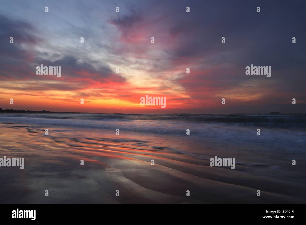 Il sole sorge in una raffica di colore arancio, rosso e viola in un cielo pieno di spesse nuvole. Le onde del mare si schiumano sulla sabbia bagnata e riflettente. Foto Stock