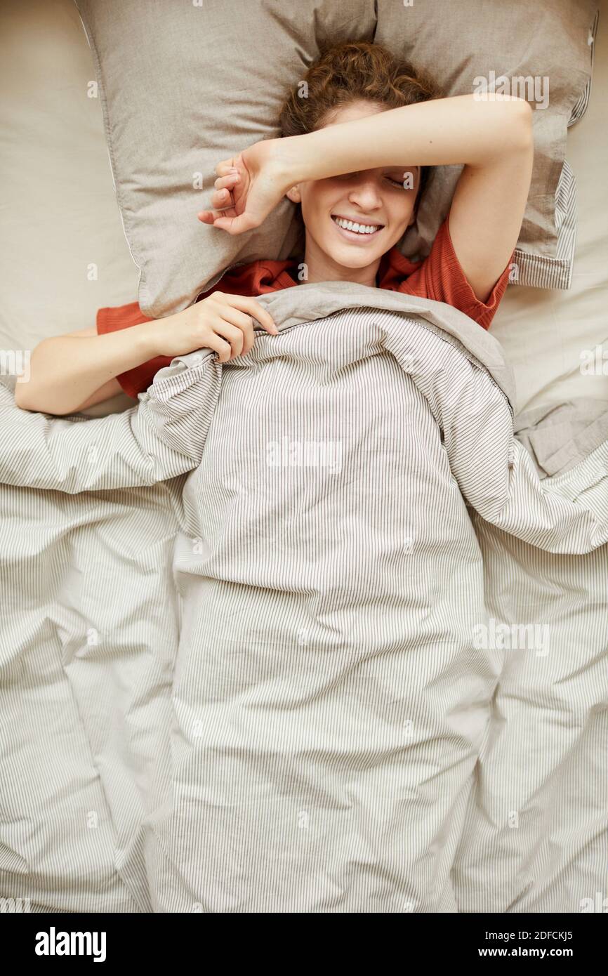 Vista ad alto angolo di una giovane donna che sorride alla fotocamera mentre riposandosi nel suo letto a casa Foto Stock