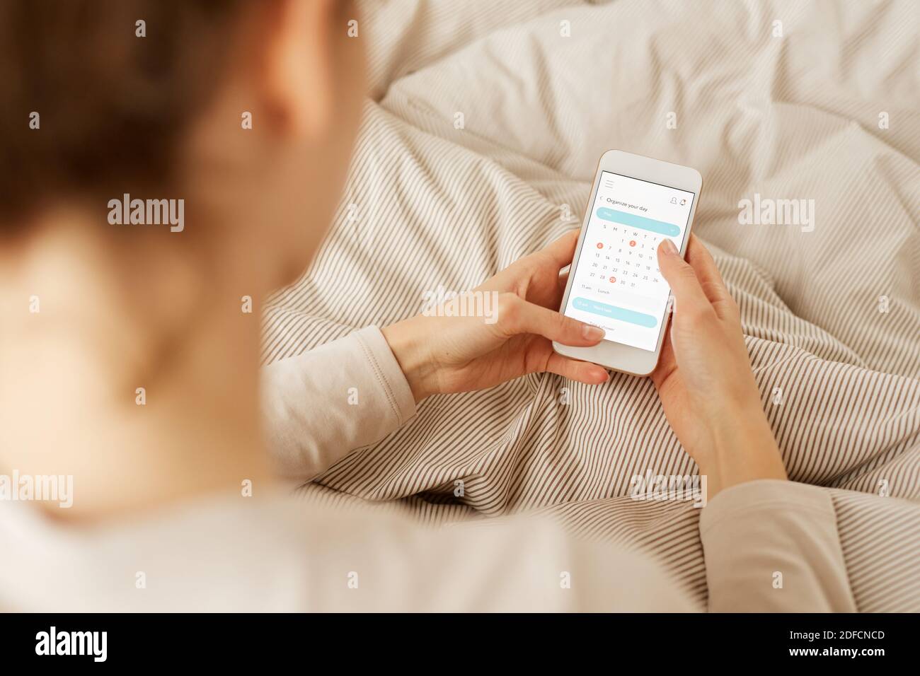 Vista posteriore della giovane donna seduta nel suo letto e. pianificare la sua giornata di lavoro in calendario online Foto Stock