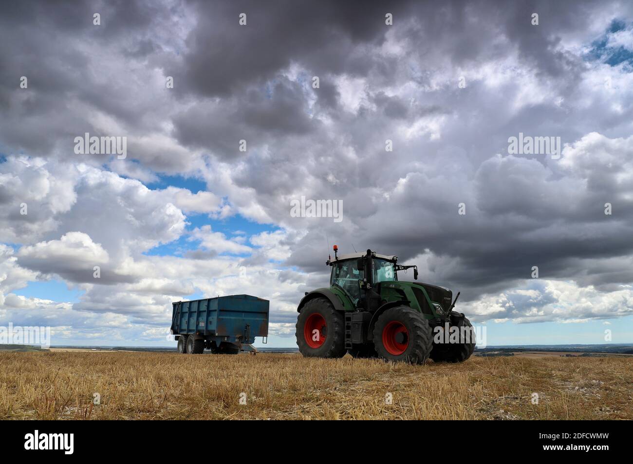 Un trattore verde e un rimorchio siedono in cima ai resti di un campo di prodotti di recente taglio sotto un cielo ampio e minaccioso riempito di precipitose grigie. Foto Stock